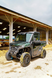 A green UFORCE U10 PRO ATV with large tires is parked in front of a farm building, hay bales and cows lounging peacefully in the background.