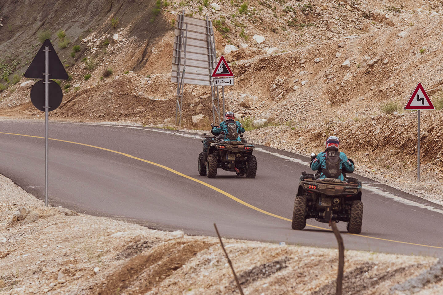 Two people ride CFMOTO ATVs on a winding road in a rocky landscape, with traffic signs visible along the route. It's as if they're part of an Albania Rally, navigating the challenging terrain like a skilled racing team.
