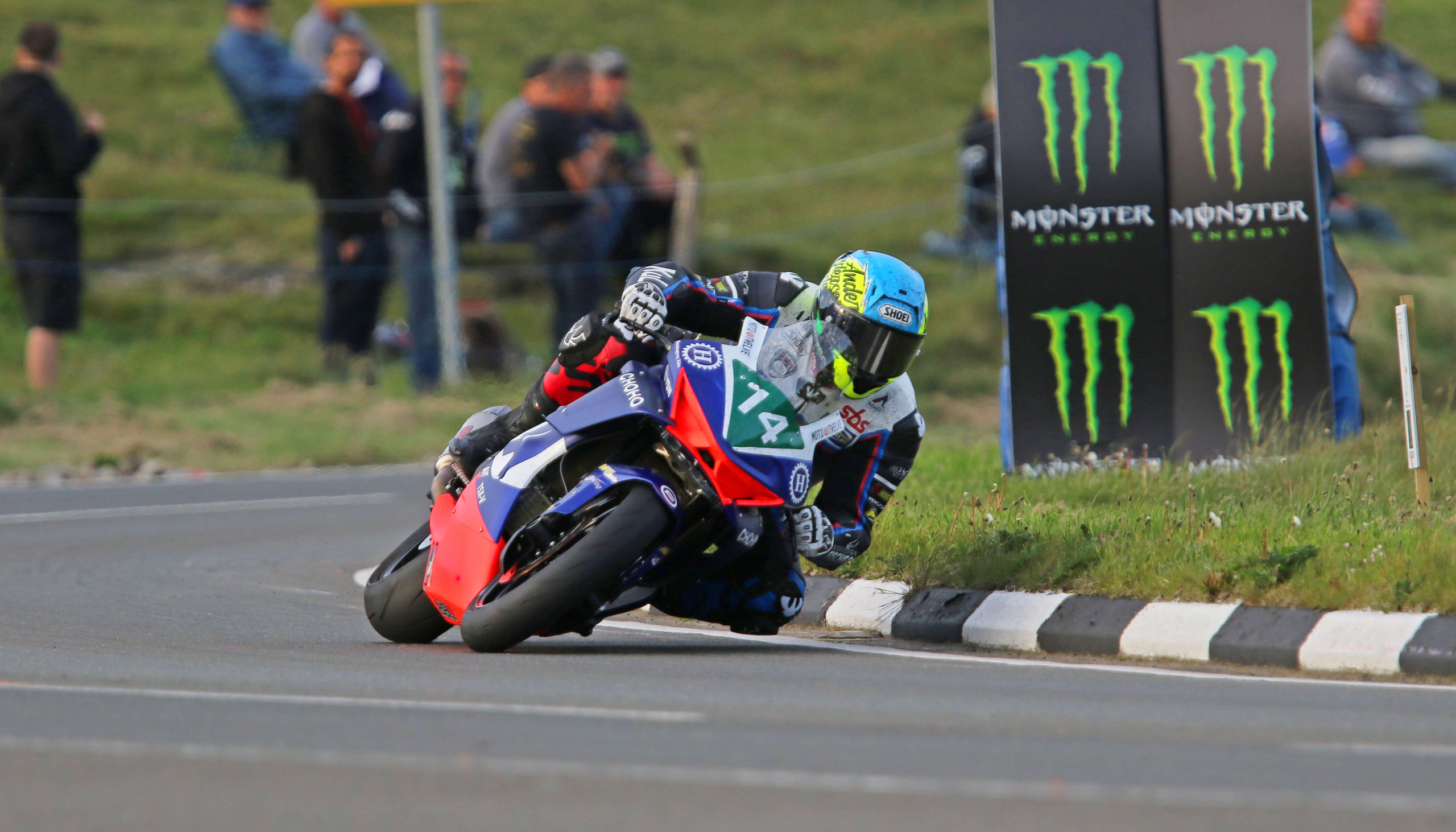 A motorcycle racer wearing number 74, competing at the Isle of Man TT, leans into a bend on a road circuit, with Monster Energy advertising hoardings and spectators in the background.