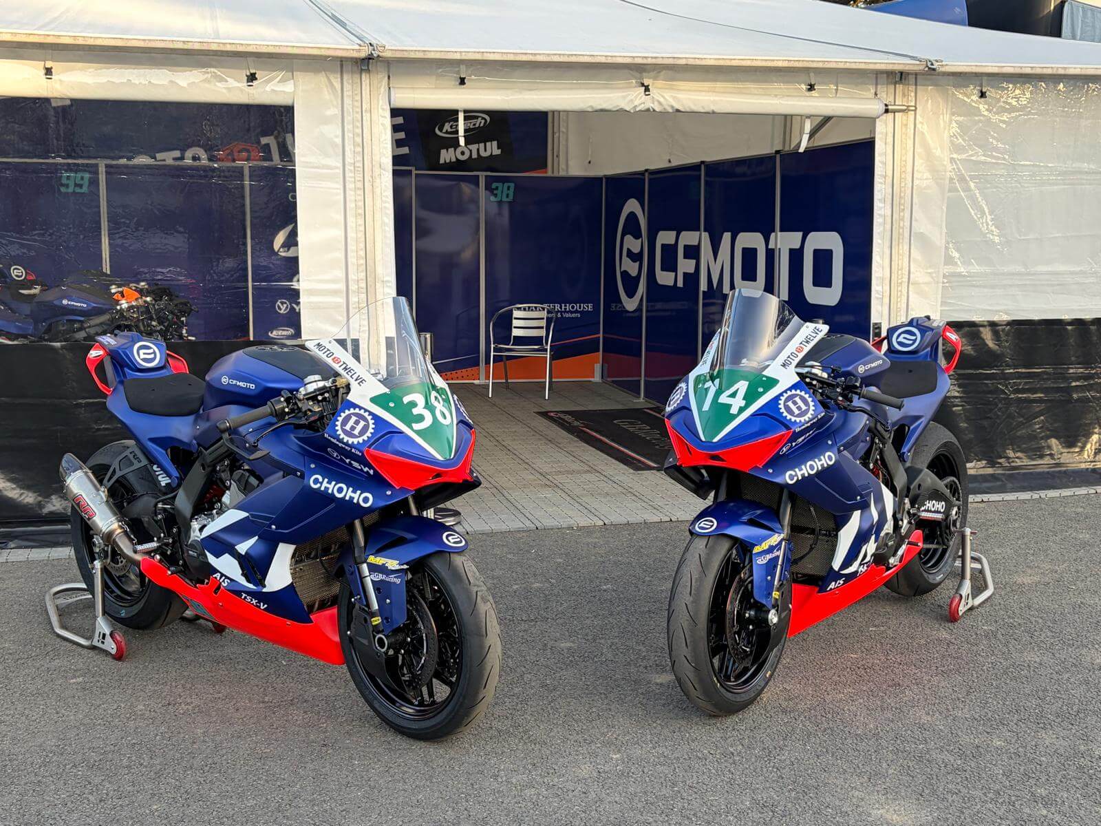 Two blue, red, and black racing motorbikes with numbers 38 and 14 are parked on stands outside a CFMOTO-branded tent at a motorsport event, showcasing the brand’s presence at the Isle of Man TT.