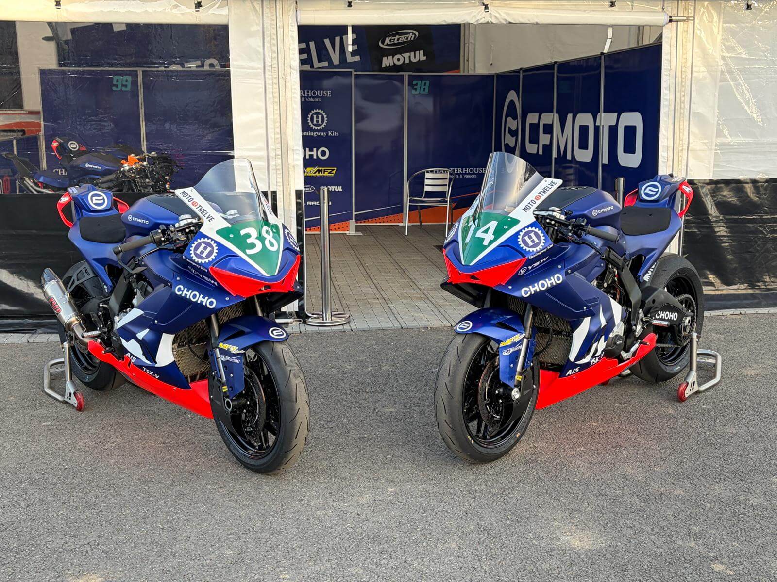 Two blue, red, and green racing motorbikes numbered 38 and 44 are parked side by side in front of a CFMOTO team garage at a motorsport event, ready to take on the excitement of Moto Twelve.