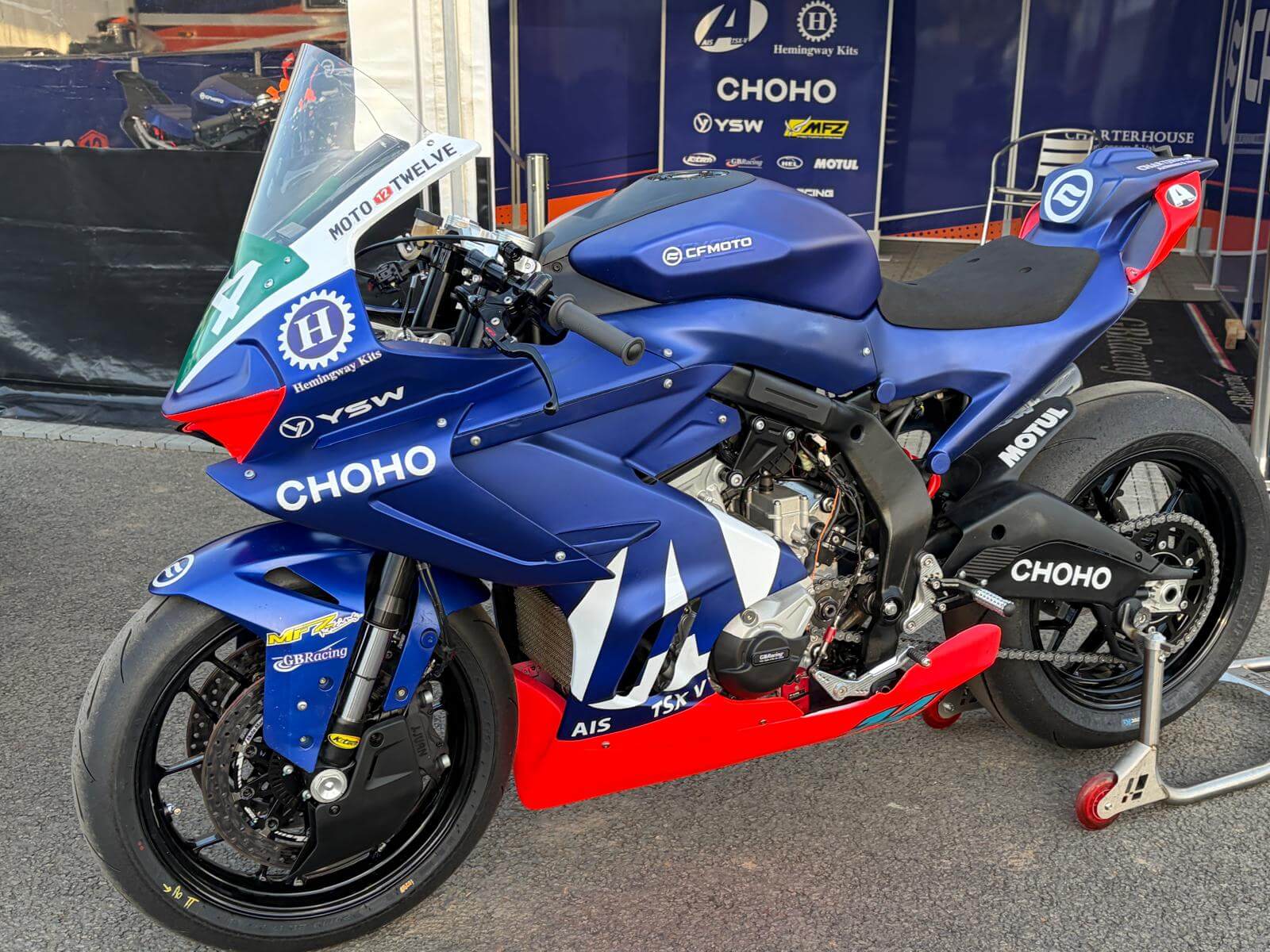 Blue CHOHO racing motorbike on paddock stand, featuring various sponsor logos and Moto Twelve decals, parked in a team pit area, ready for the Isle of Man TT.