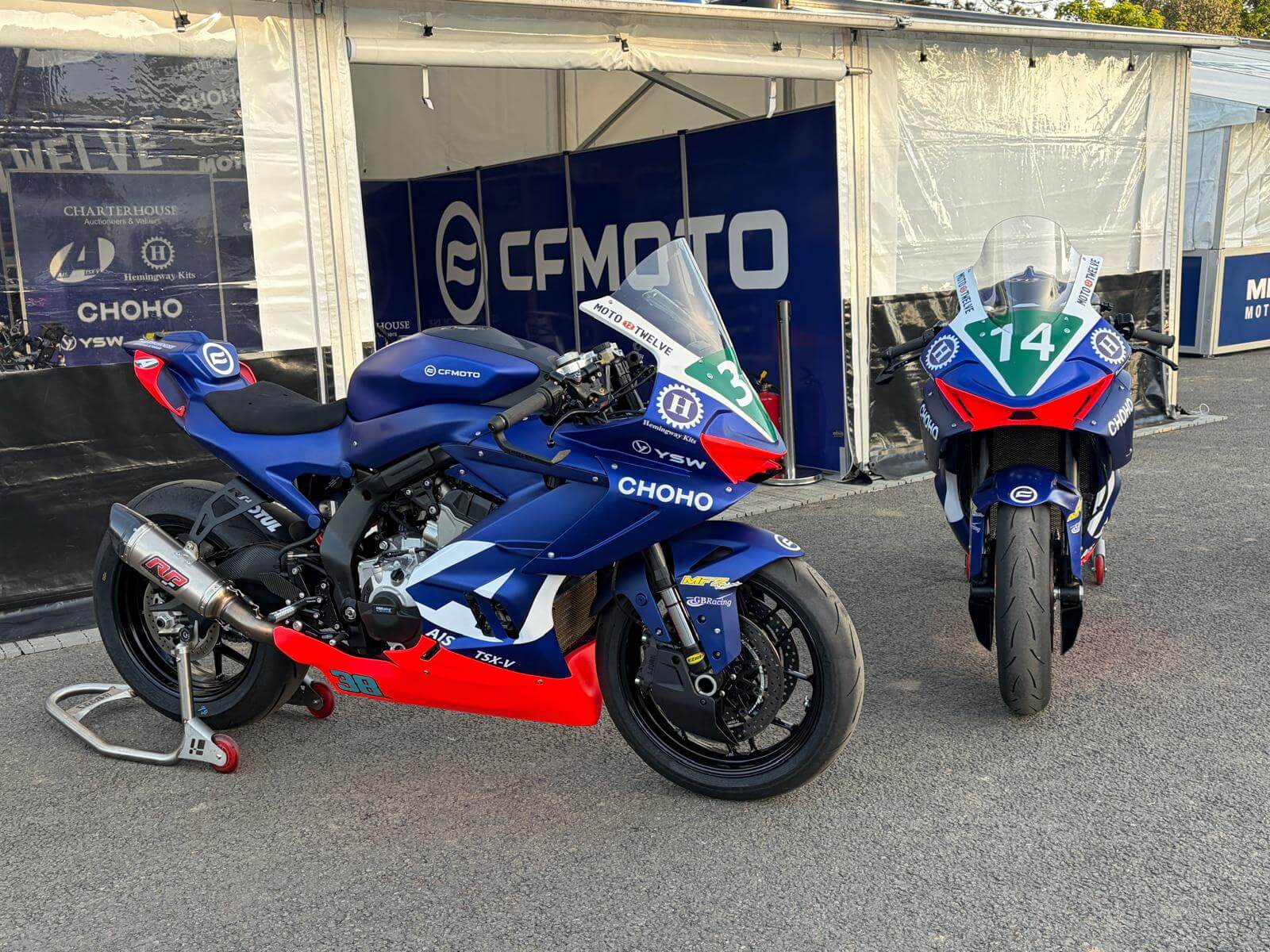 Two blue and red CFMOTO racing motorbikes are parked on tarmac outside a tent with CFMOTO branding, ready for the next Moto Twelve event at the Isle of Man TT.