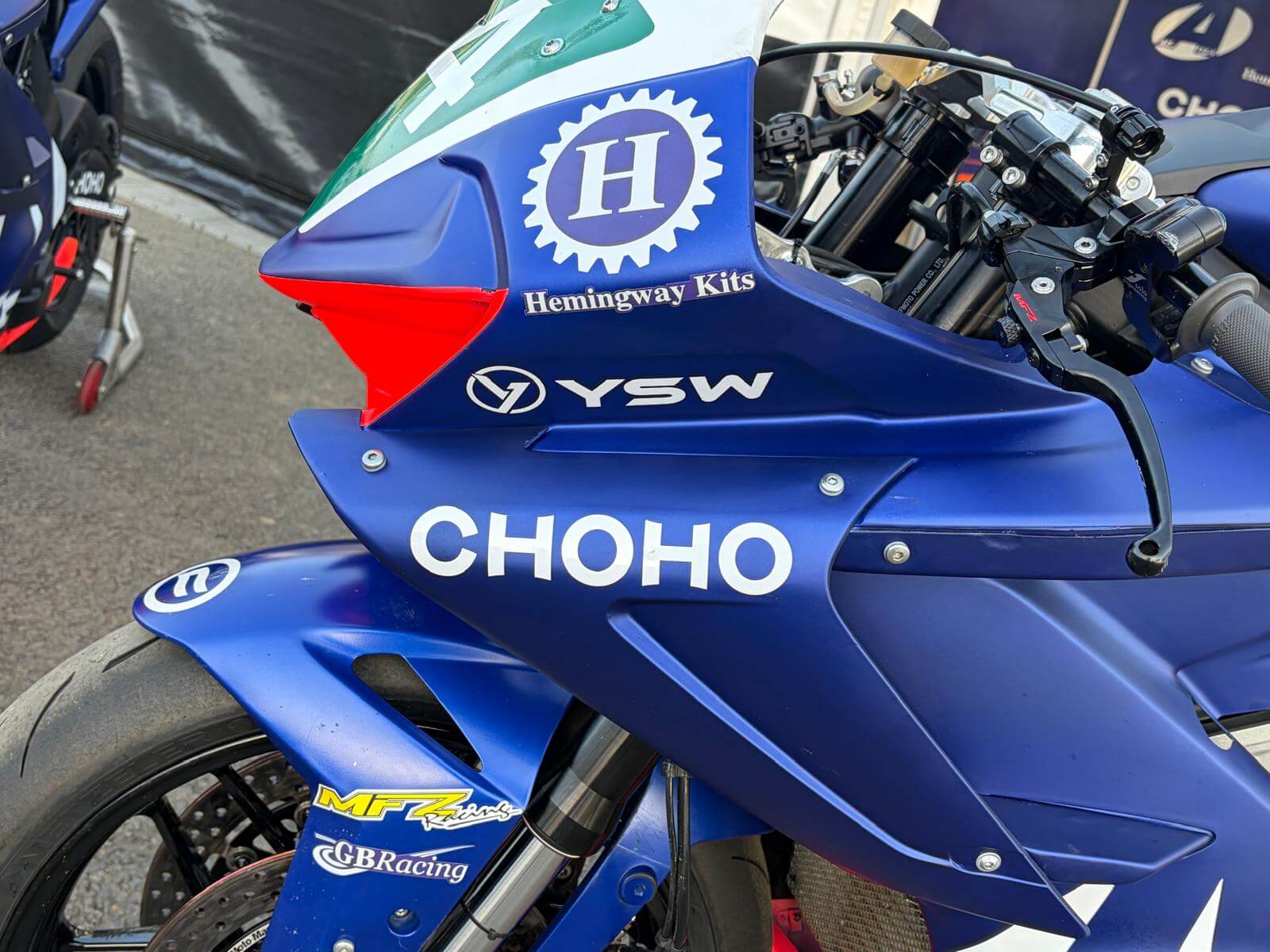 Close-up of a blue racing motorbike, possibly a Moto Twelve or CFMOTO, showing sponsor logos like "Hemingway Kits," "YSW," "CHOHO," and "MFZ Racing." The bike is parked on a paved surface, evoking the spirit of the Isle of Man TT.