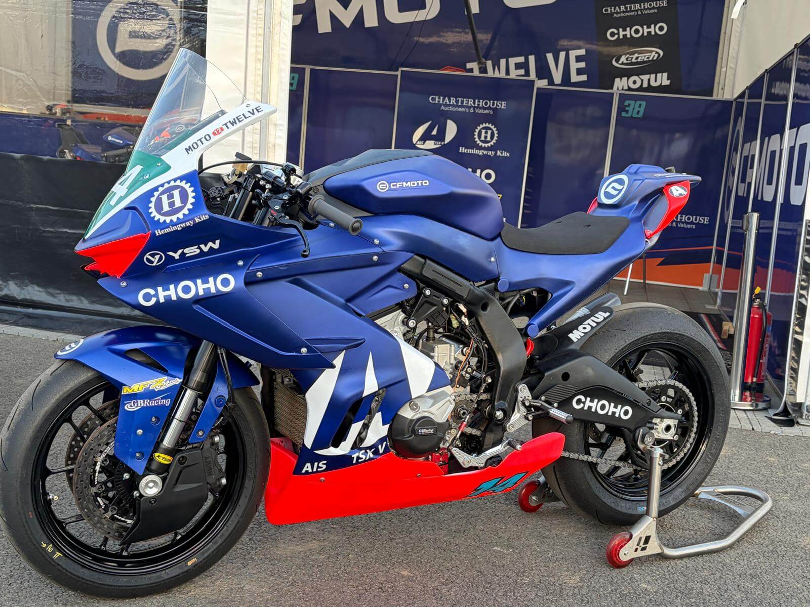 A blue CFMOTO racing motorbike with Moto Twelve, CHOHO, and YSW branding is parked on a paddock stand in a team pit area, ready for action at the Isle of Man TT.