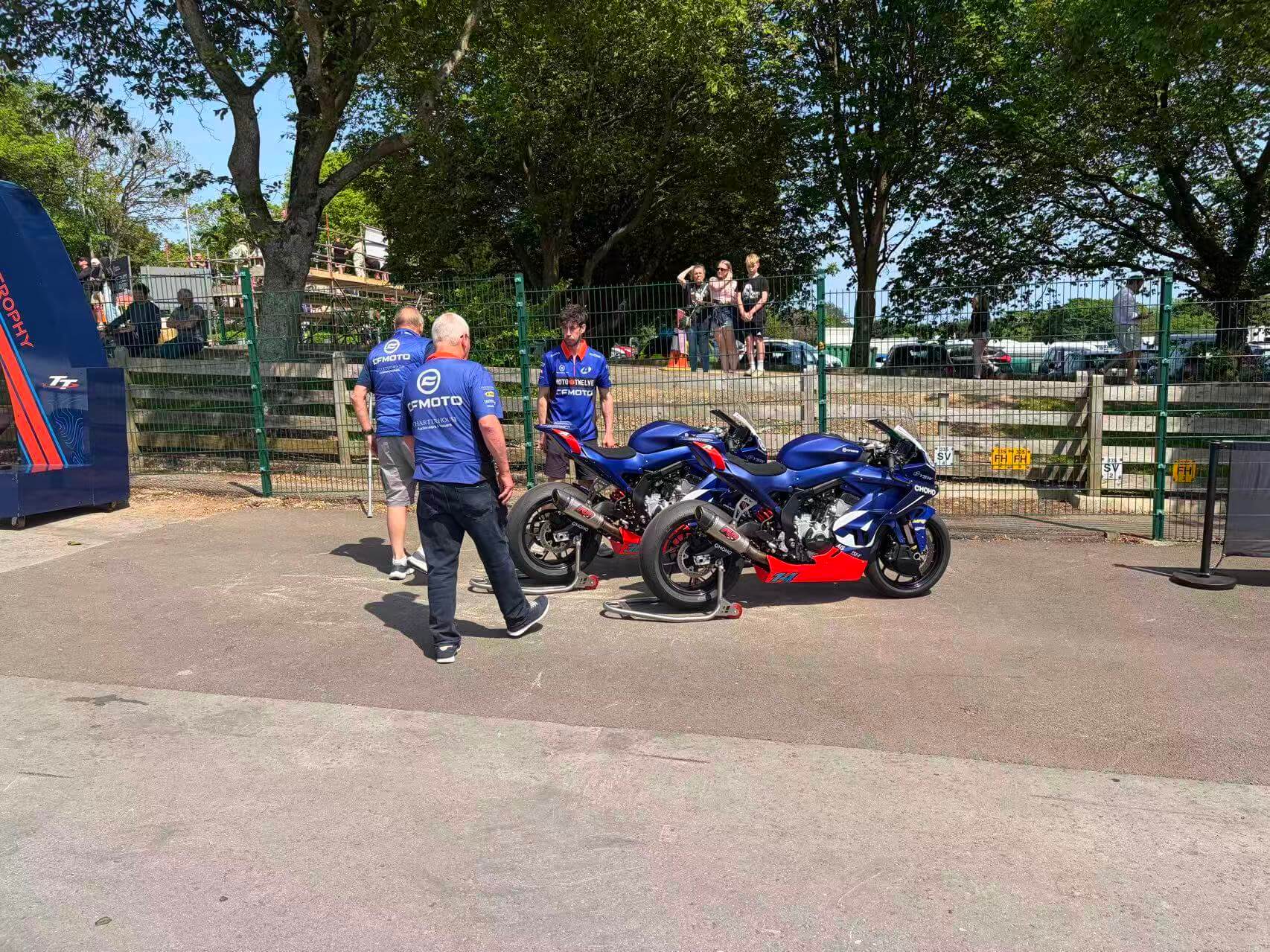 Several people stand near two blue CFMOTO motorbikes parked on stands in an outdoor area, with trees and a fence in the background.