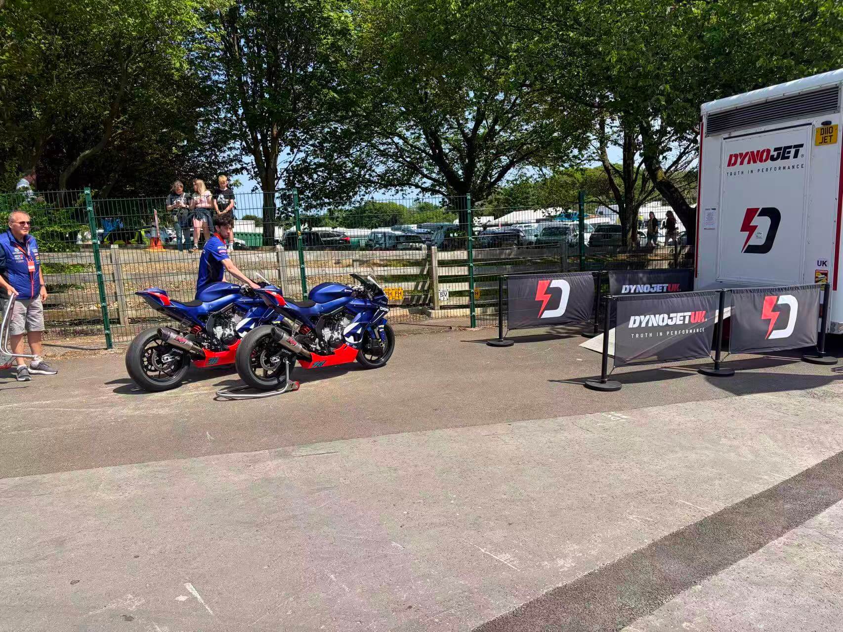 A person sits on a blue CFMOTO motorbike positioned next to a Dynojet testing station outdoors, with trees and parked cars in the background.