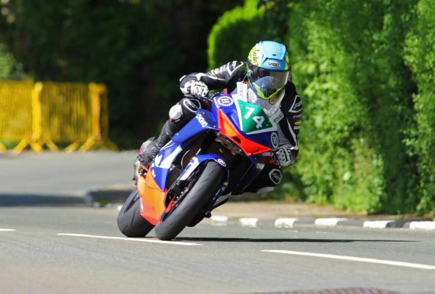 A motorcyclist wearing racing kit and a blue helmet rides a Moto Twelve sport bike numbered 74 on a tarmac road, leaning into a bend with greenery in the background, reminiscent of the Isle of Man TT.
