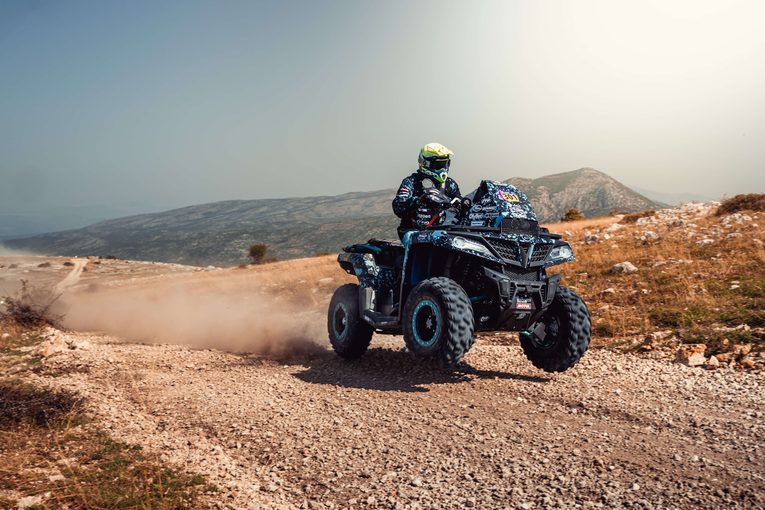 A member of the Thunder Racing Team is riding an ATV quickly on a dusty, rocky trail through a mountainous landscape in Portugal under a clear sky.