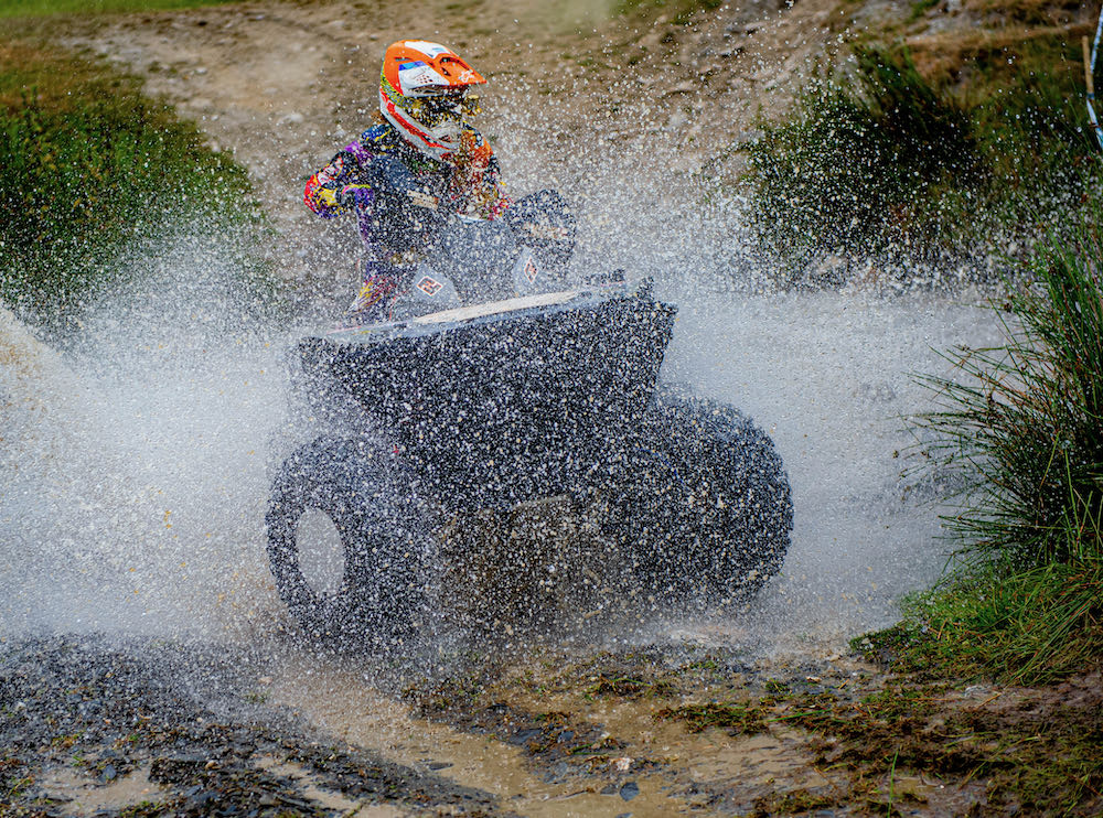 On a lively weekend, a person races an ATV through a muddy puddle, sending splashes skyward against the backdrop of Wales' lush grassy terrain.