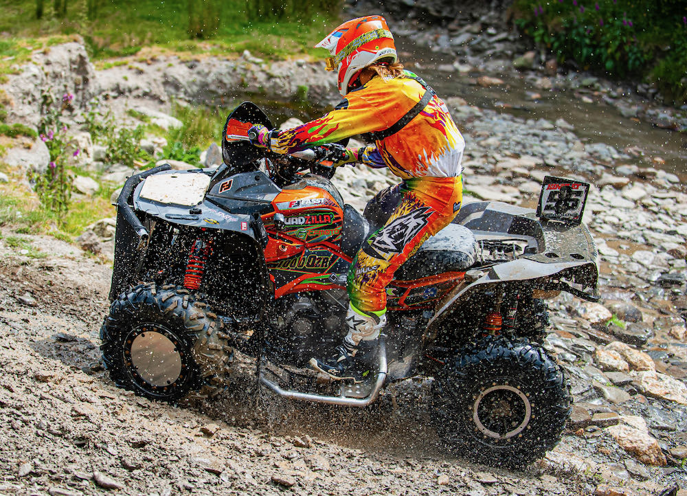 A person confidently rides their ATV up a rocky hill in vibrant orange and yellow racing gear, helmet securely fastened, as water splashes from the wheels during a thrilling weekend ATV race in Wales.