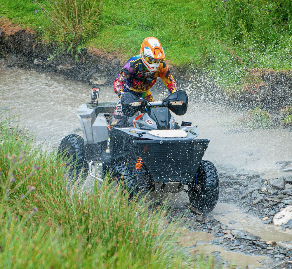 Revving up for a thrilling weekend, a person maneuvers their ATV through a muddy water trail in Wales, creating spectacular splashes all around.