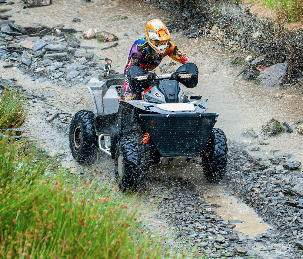 A person navigates a rugged Welsh landscape during an exhilarating ATV race, kicking up splashes of mud and water as they conquer the challenging terrain.