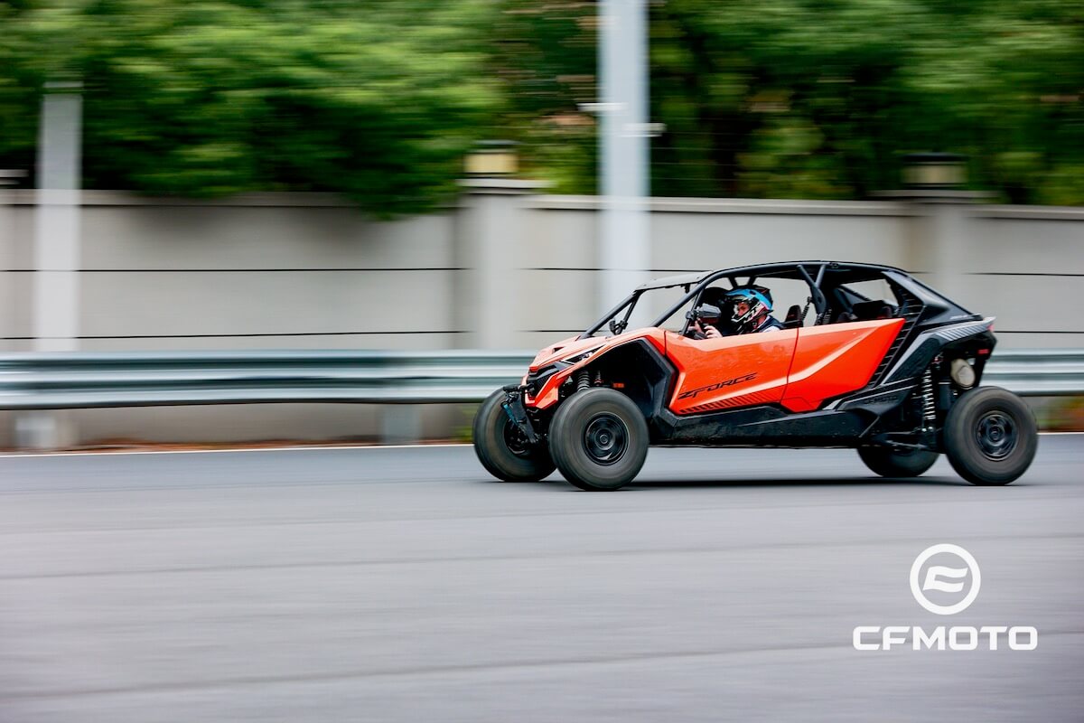 A red and black off-road buggy, one of the CFMOTO classics, speeds down a paved road, its blurred background conveying pure motion. The CFMOTO logo is visible in the bottom right corner.