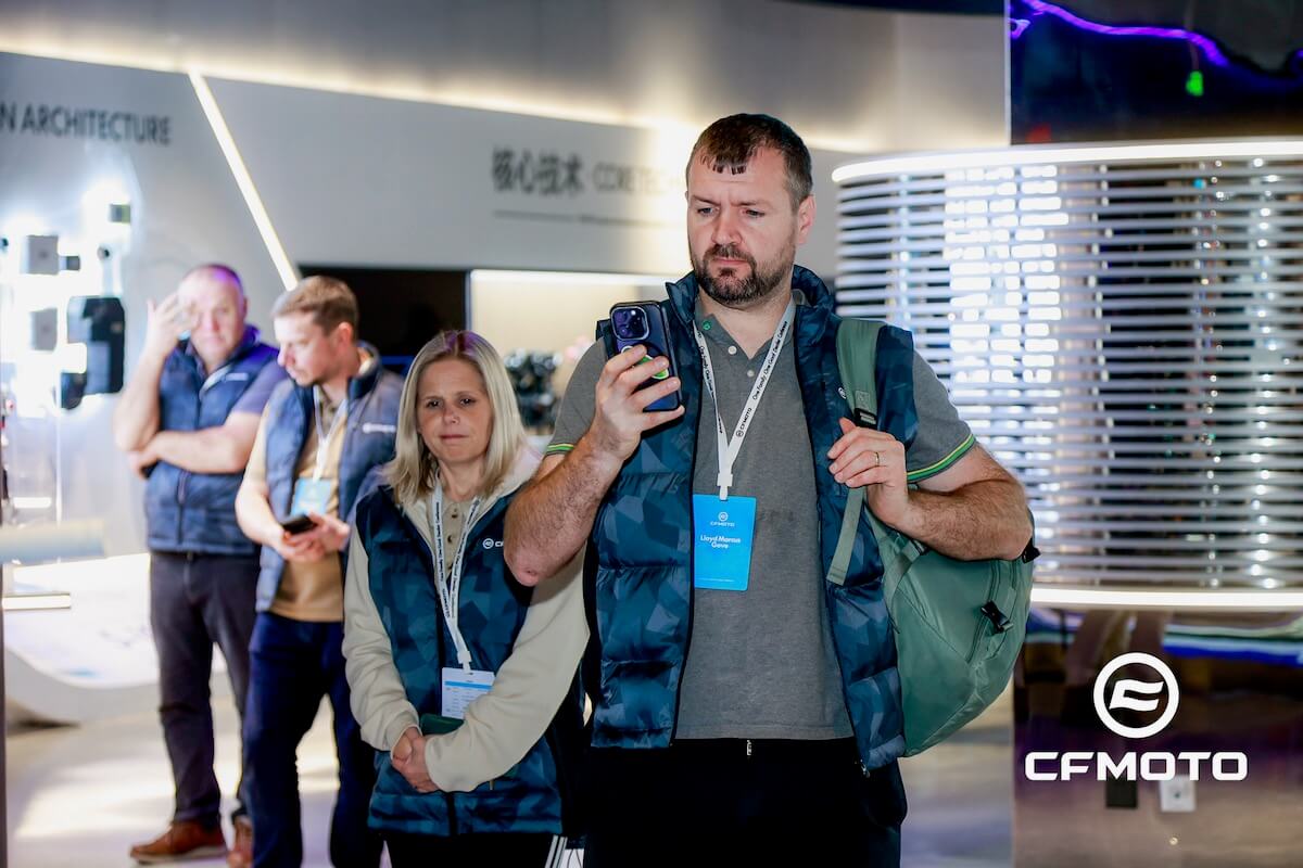 A group of people wearing blue vests stand indoors; one man is holding a smartphone, and the CFMOTO logo—known for CFMOTO classics—is visible in the bottom right corner.