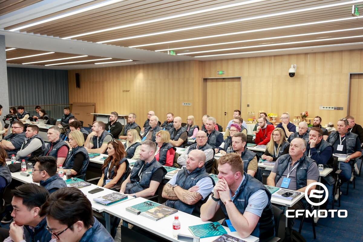 A large group of people sit at tables in a modern conference room, listening attentively; the CFMOTO logo in the bottom right hints at a presentation on CFMOTO classics.