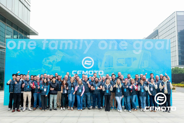 A large group of people pose together outside in front of a blue CFMOTO banner that reads “one family one goal,” with most giving a thumbs up, celebrating their shared passion for CFMOTO classics.