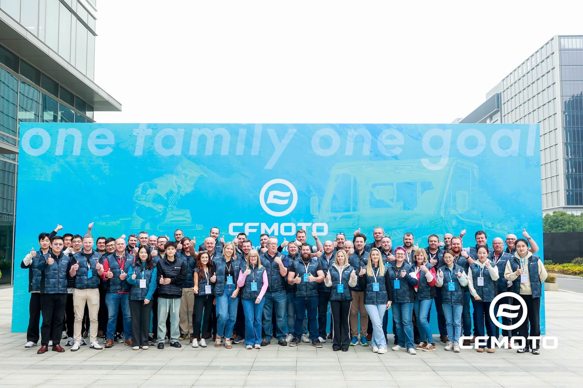 A large group of people pose together outside in front of a blue CFMOTO banner that reads “one family one goal,” with most giving a thumbs up, celebrating their shared passion for CFMOTO classics.
