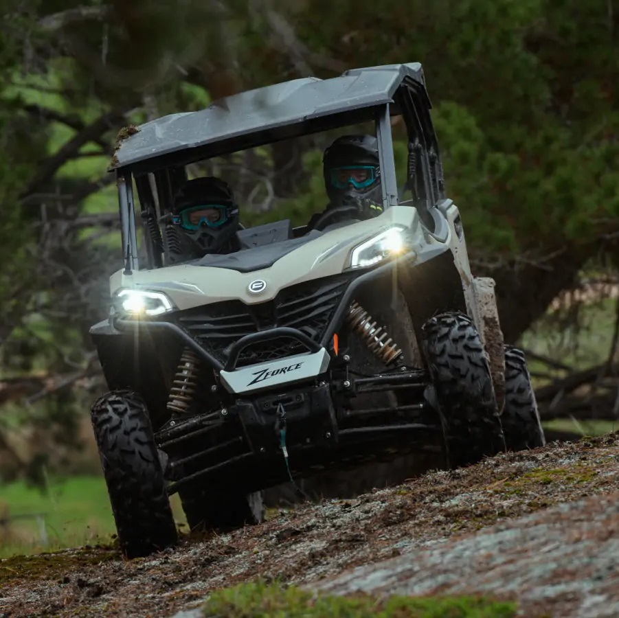 Two people drive an off-road vehicle on a rocky path surrounded by trees.