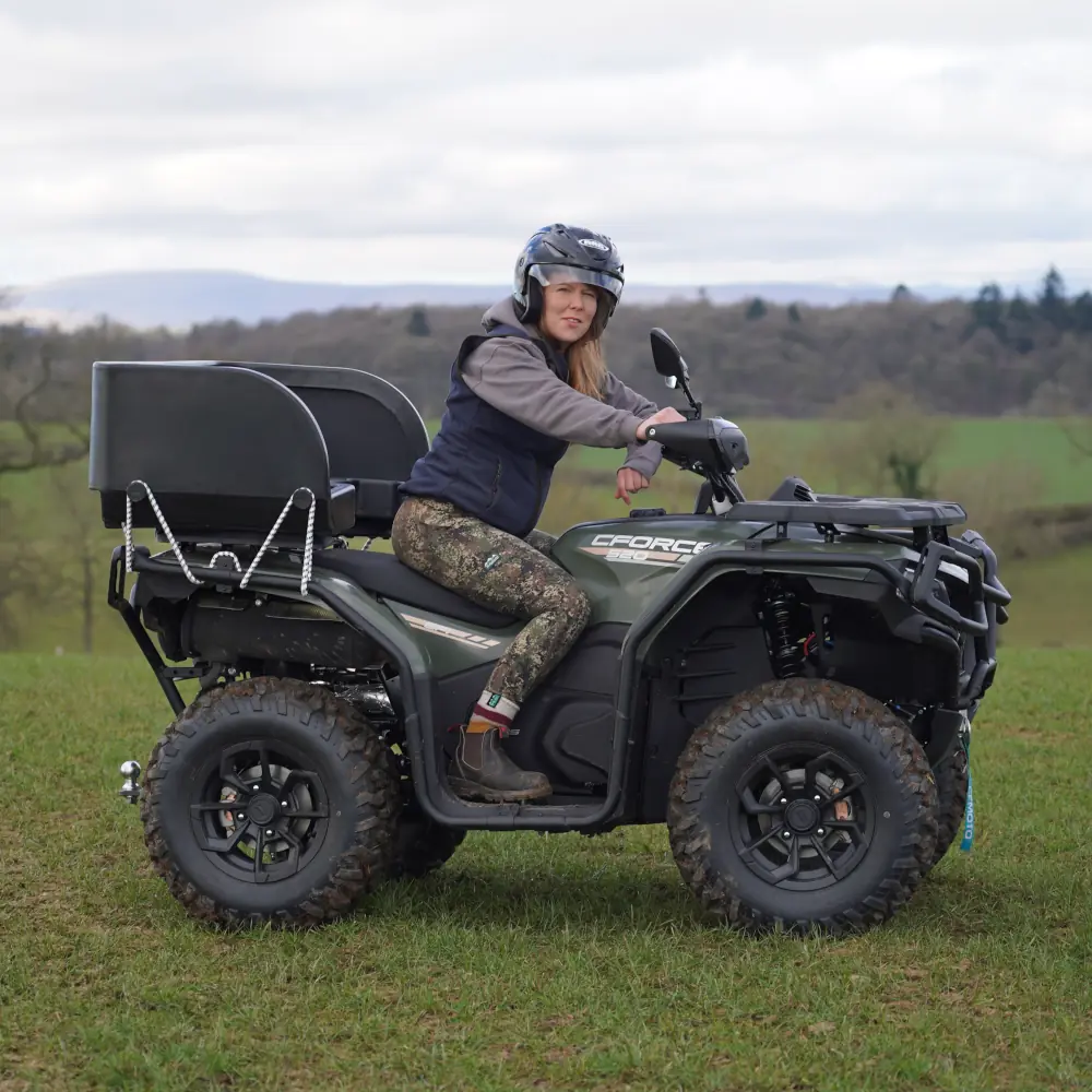 A person wearing a helmet and outdoor clothing sits on a CForce 520 all-terrain vehicle parked on a grassy field, with trees in the background.