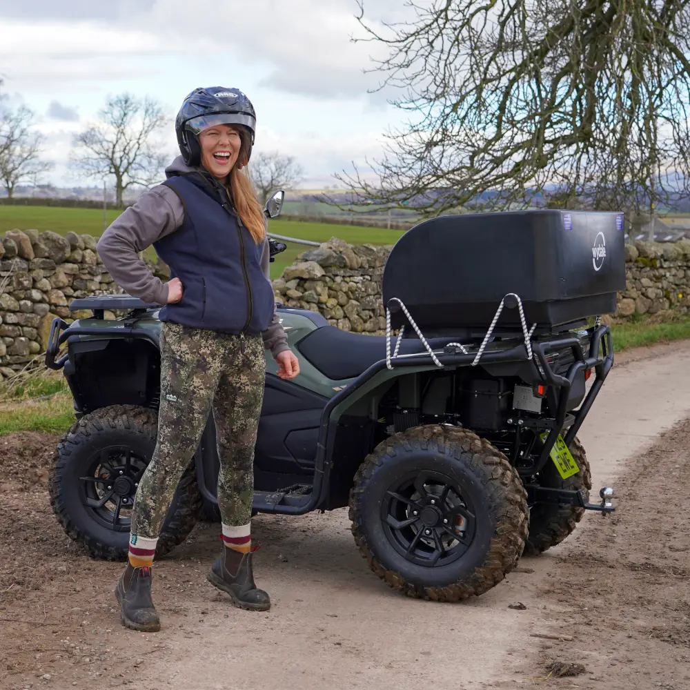 A woman in a helmet and outdoor clothing stands next to a CForce 520 ATV on a dirt road, with a stone wall and fields in the background.