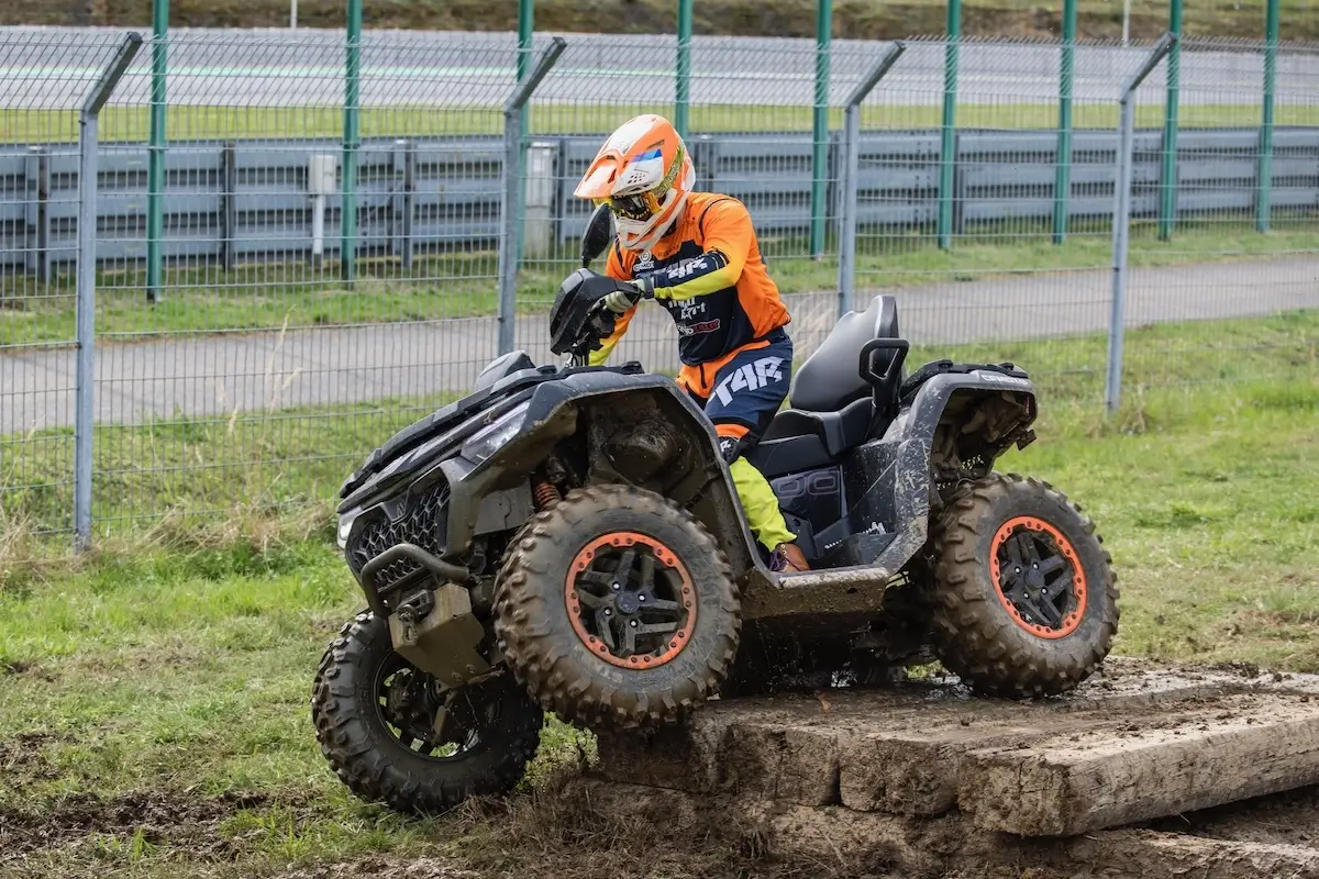 A person wearing orange and black gear rides a CForce 1000 Touring Premium ATV over a muddy wooden obstacle on a grassy off-road track with metal fences in the background.