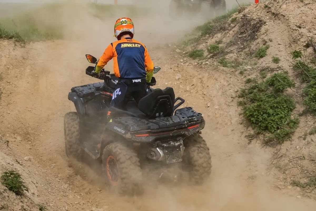 Wearing a helmet and protective gear, a person rides a CForce 1000 Touring Premium ATV along a dusty dirt trail surrounded by patches of grass and small hills.