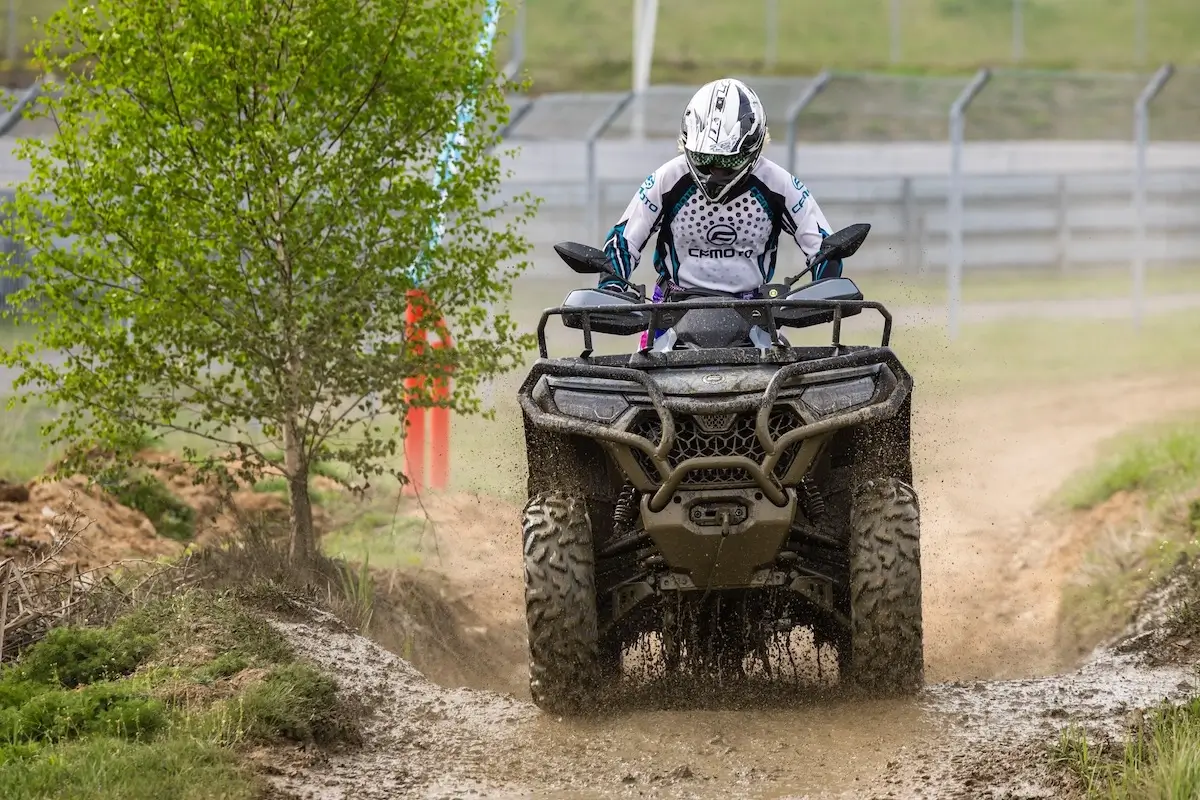 A person wearing a helmet and protective gear rides a CForce 850 Touring ATV on a dirt track, with mud splashing from the wheels and a small tree nearby.