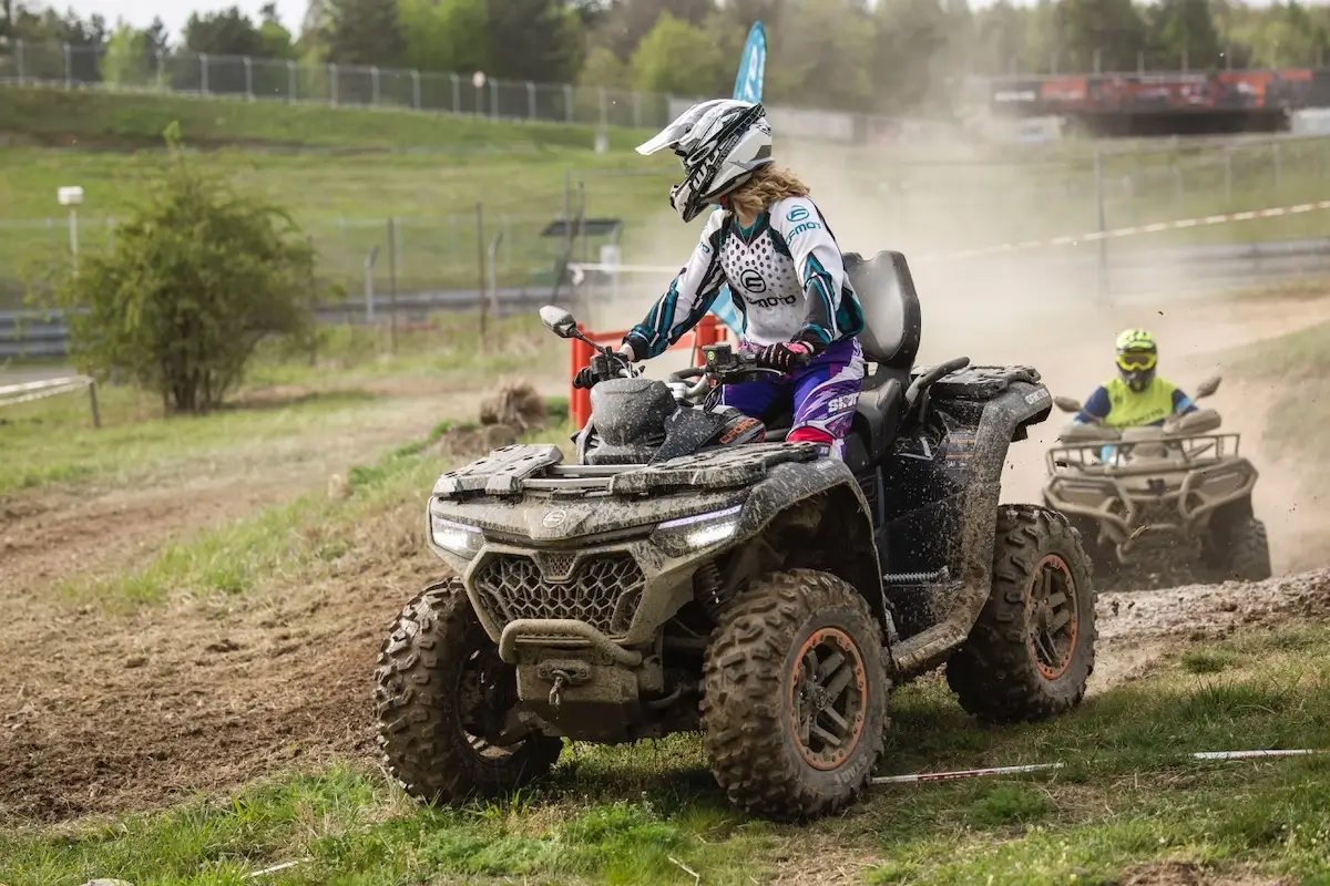 A person rides a CForce 1000 Touring Premium all-terrain vehicle on a dirt track, wearing a helmet and protective gear, with another rider visible in the background.