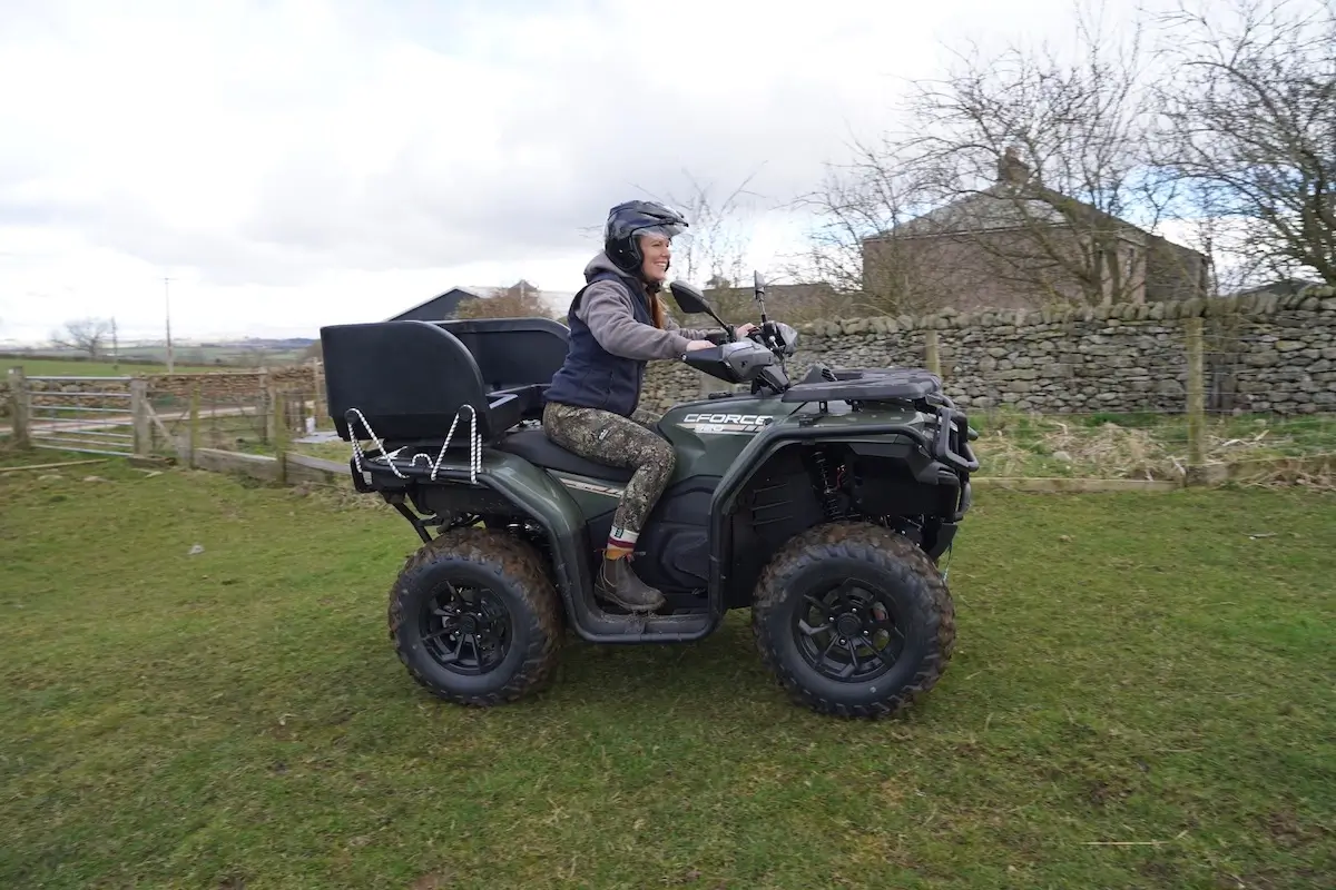 A person wearing a helmet and outdoor clothing rides a CForce 520 ATV across a grassy area, with a stone wall and house visible in the background.