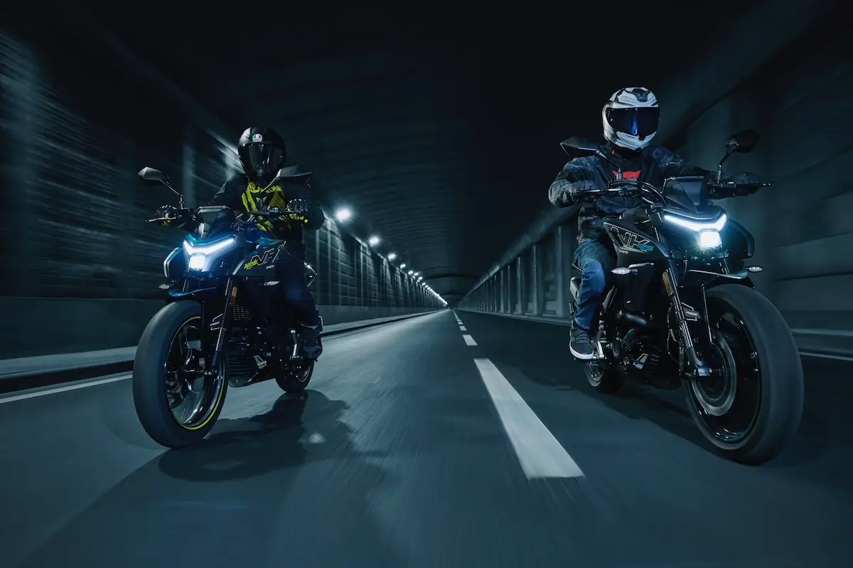 Two motorcyclists ride side by side on a 125NK inside a dimly lit tunnel, both wearing helmets and protective gear.