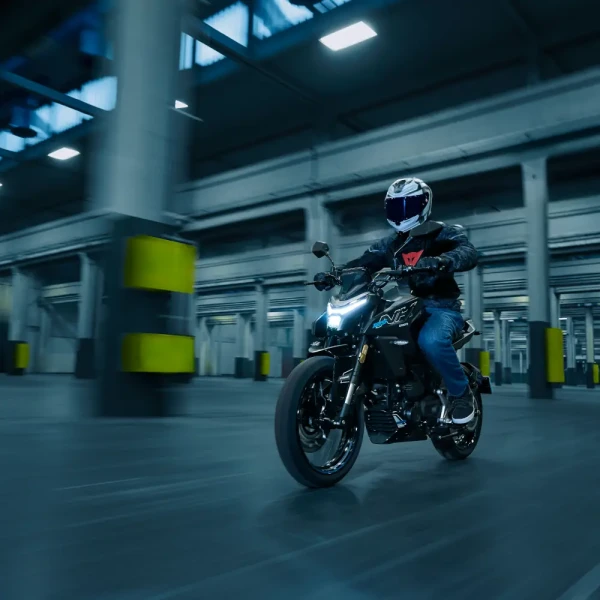 A person wearing a helmet rides a black motorcycle through a large, industrial indoor space with concrete pillars, making it an ideal setting for new riders to practice their skills.