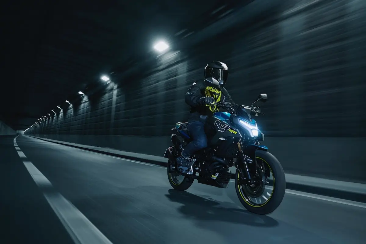 A person wearing a helmet and jacket rides a blue 125NK motorcycle at night through a well-lit tunnel with empty lanes.