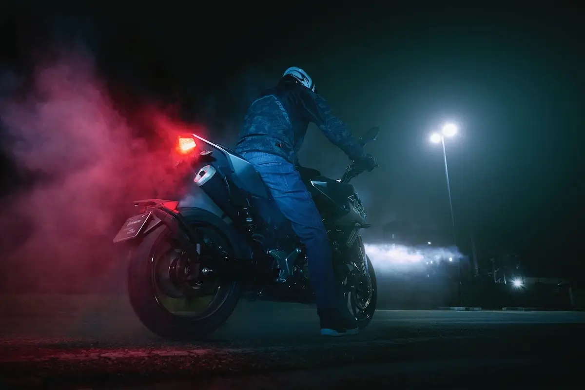 A person wearing a helmet and jacket sits on a 125NK motorcycle at night, with bright lights and colored smoke in the background.