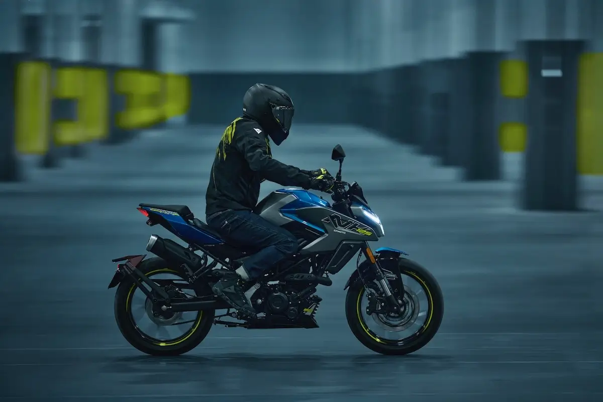 A person in full black riding gear and helmet rides a blue and black 125NK motorcycle in an indoor, industrial-looking space with yellow markings on pillars.