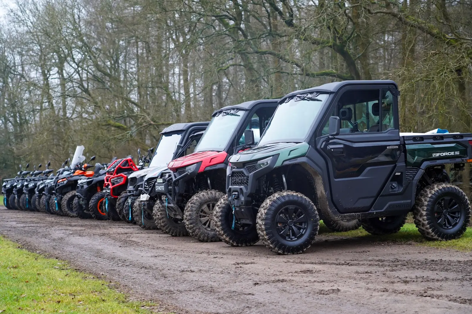 A row of off-road vehicles and ATVs, including a farm quad, is parked along a dirt path beside a grassy area, with trees in the background.