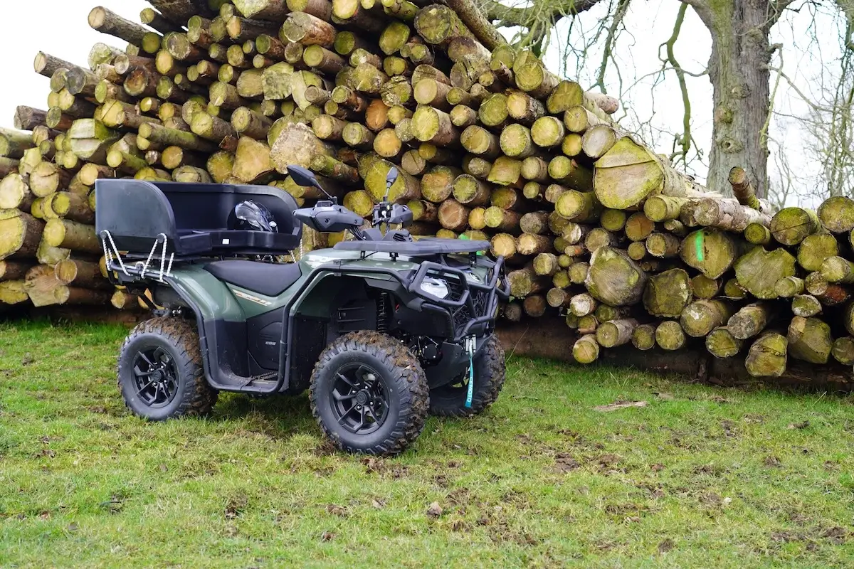 A green CForce 520 quad bike with a rear storage box is parked on grass in front of a large stack of cut logs.