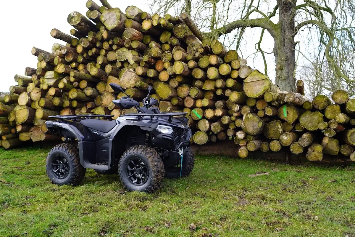 A black CForce 450 ATV is parked on grass in front of a large stack of cut logs, with a leafless tree visible in the background.