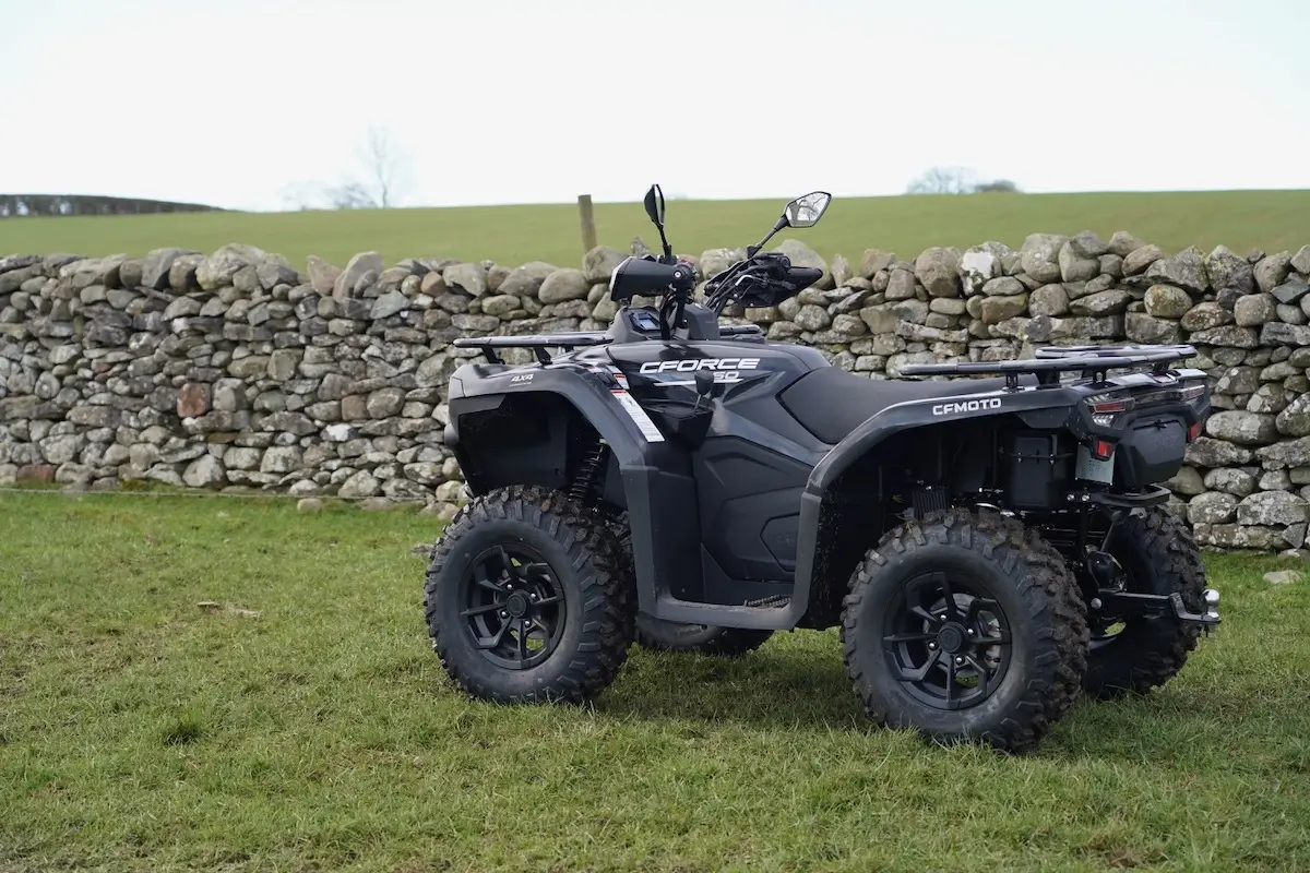 A black CFMOTO CFORCE 450 all-terrain vehicle is parked on grass near a stone wall, with a field and trees in the background.