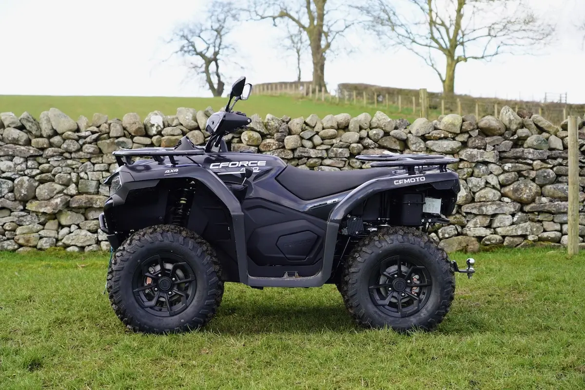 A black CFORCE CFMOTO cforce 450 all-terrain vehicle is parked on grass in front of a stone wall with bare trees in the background.