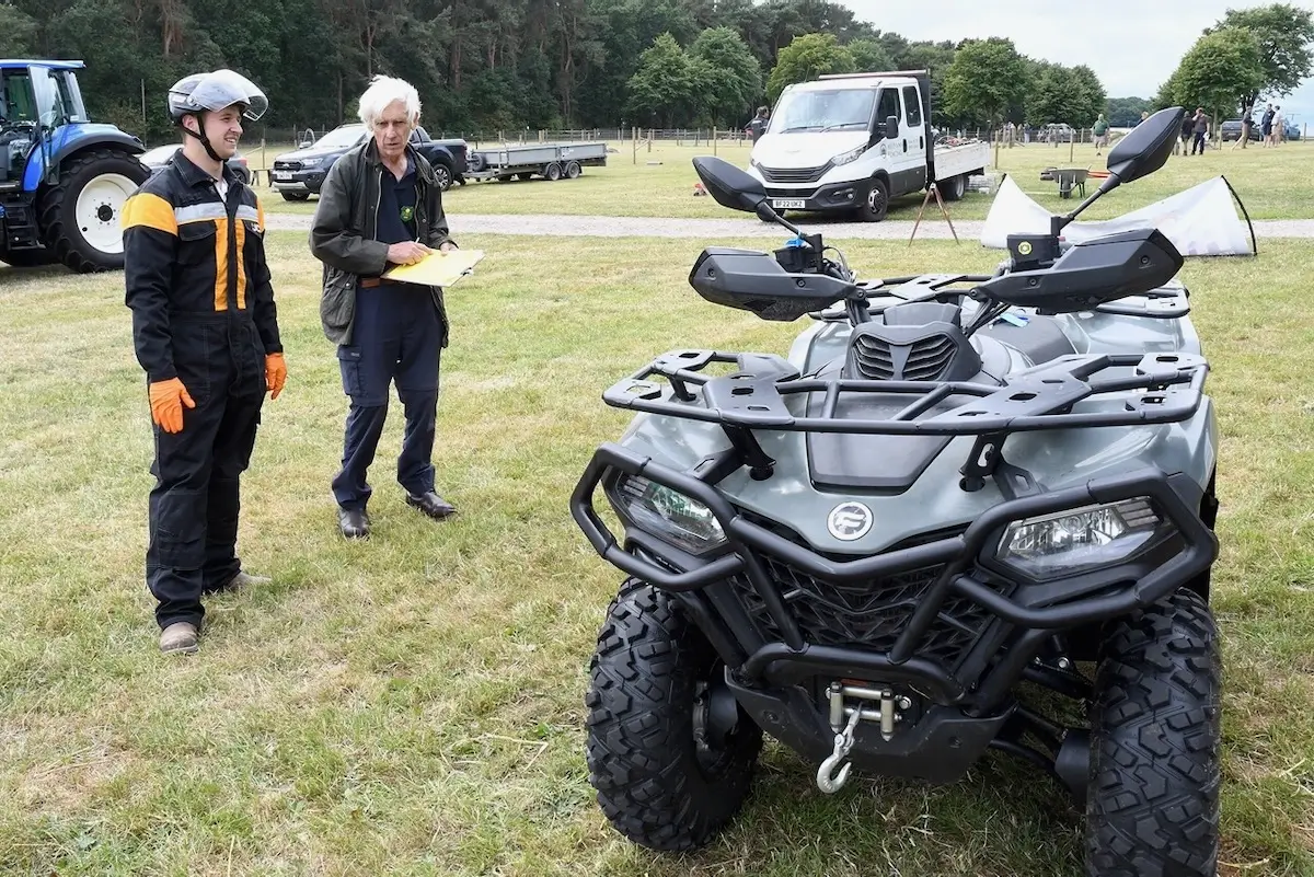Two people stand on grass near an all-terrain vehicle (ATV); one holds a clipboard, and various trucks, UTVs, and trees are visible in the background.