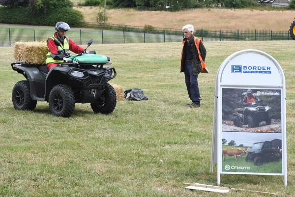 A person on an ATV with a bale of hay on the back faces another person standing nearby; a promotional sign for CFMOTO ATVs highlights their role in modern agriculture.