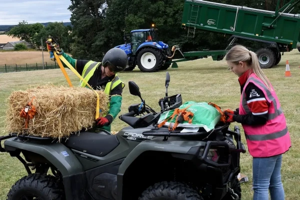 Two people secure a hay bale and a green tarp onto an ATV in a grassy field, showcasing common agriculture practices. A tractor and trailer are visible in the background.