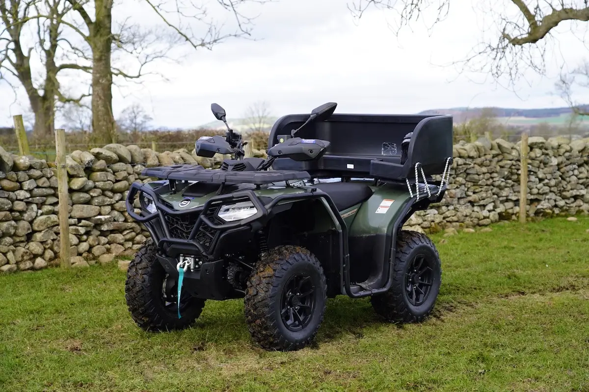A green CForce 520 ATV with a large cargo box attached to the rear is parked on grass near a stone wall and leafless trees.