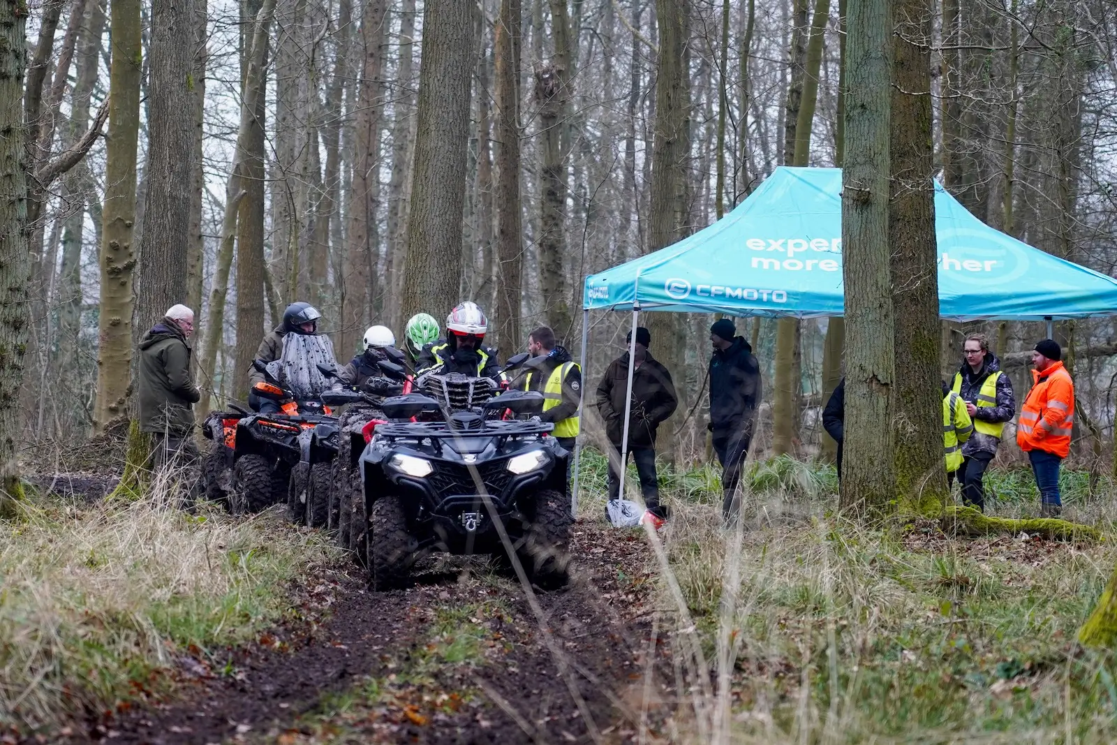 ATVs and farm quads navigate a forest trail near a blue canopy, surrounded by people in safety vests and jackets.
