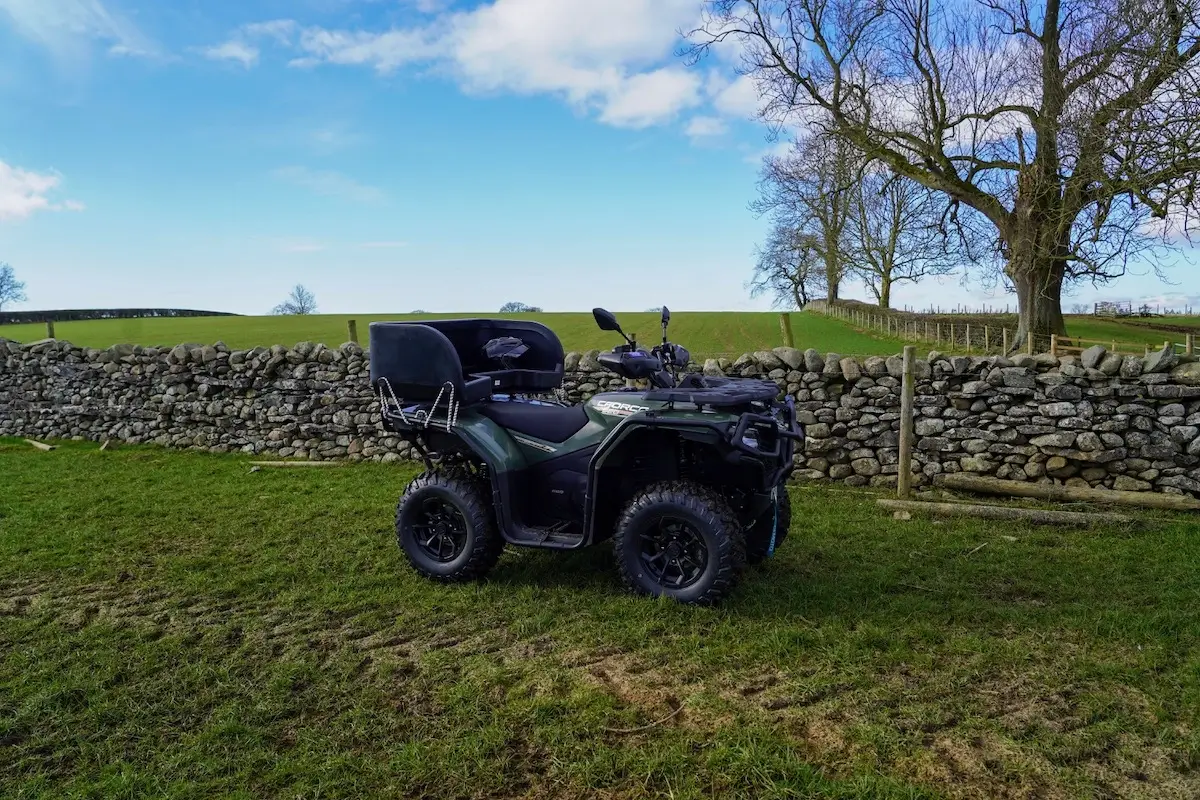 A black CForce 520 all-terrain vehicle is parked on grass near a stone wall, with leafless trees and a field in the background under a blue sky.