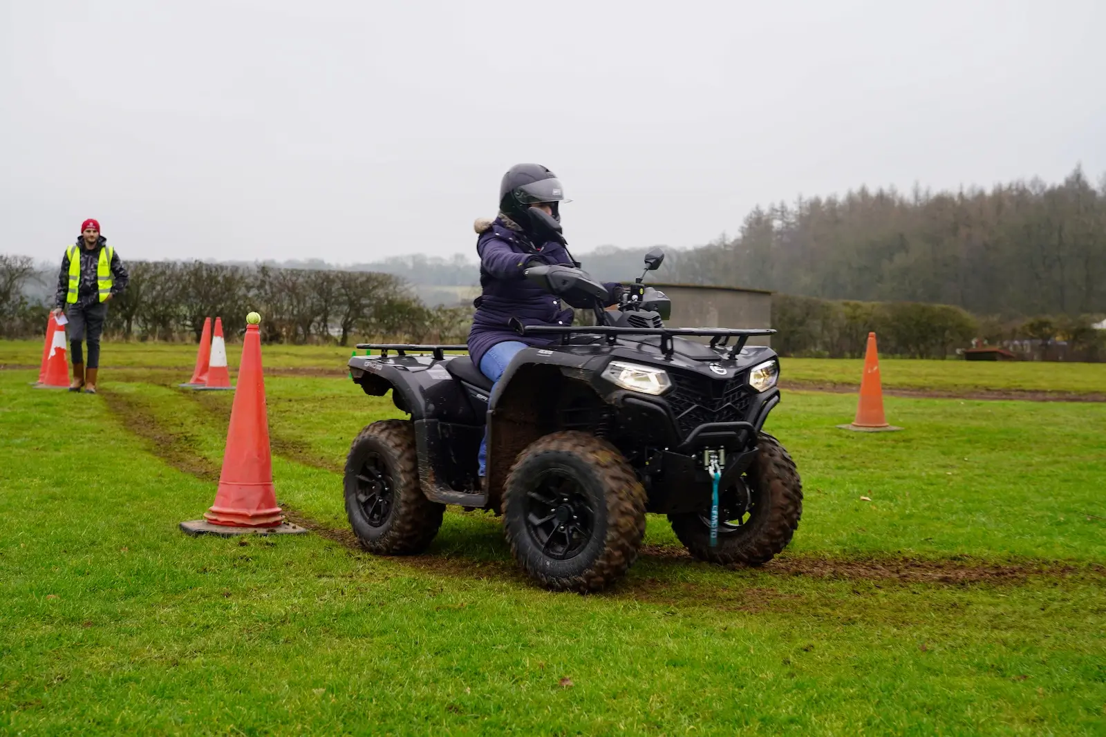 A person in protective gear rides a farm quad on the grass, skillfully navigating around orange cones. Another individual in a high-visibility jacket observes from a distance.