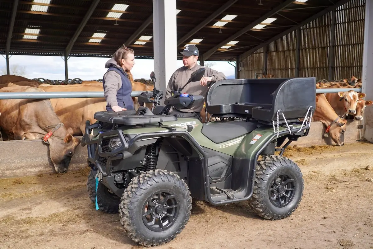 Two people stand next to a CFMoto CForce 520 ATV in a barn, talking, while several cows feed from a trough in the background.