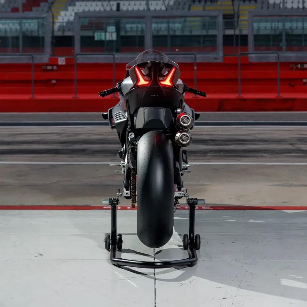 A rear view of a black CFMOTO CFORCE TECH GEN 4 Concept racing motorcycle on a stand, positioned in a pit lane at a racetrack with empty grandstands in the background.