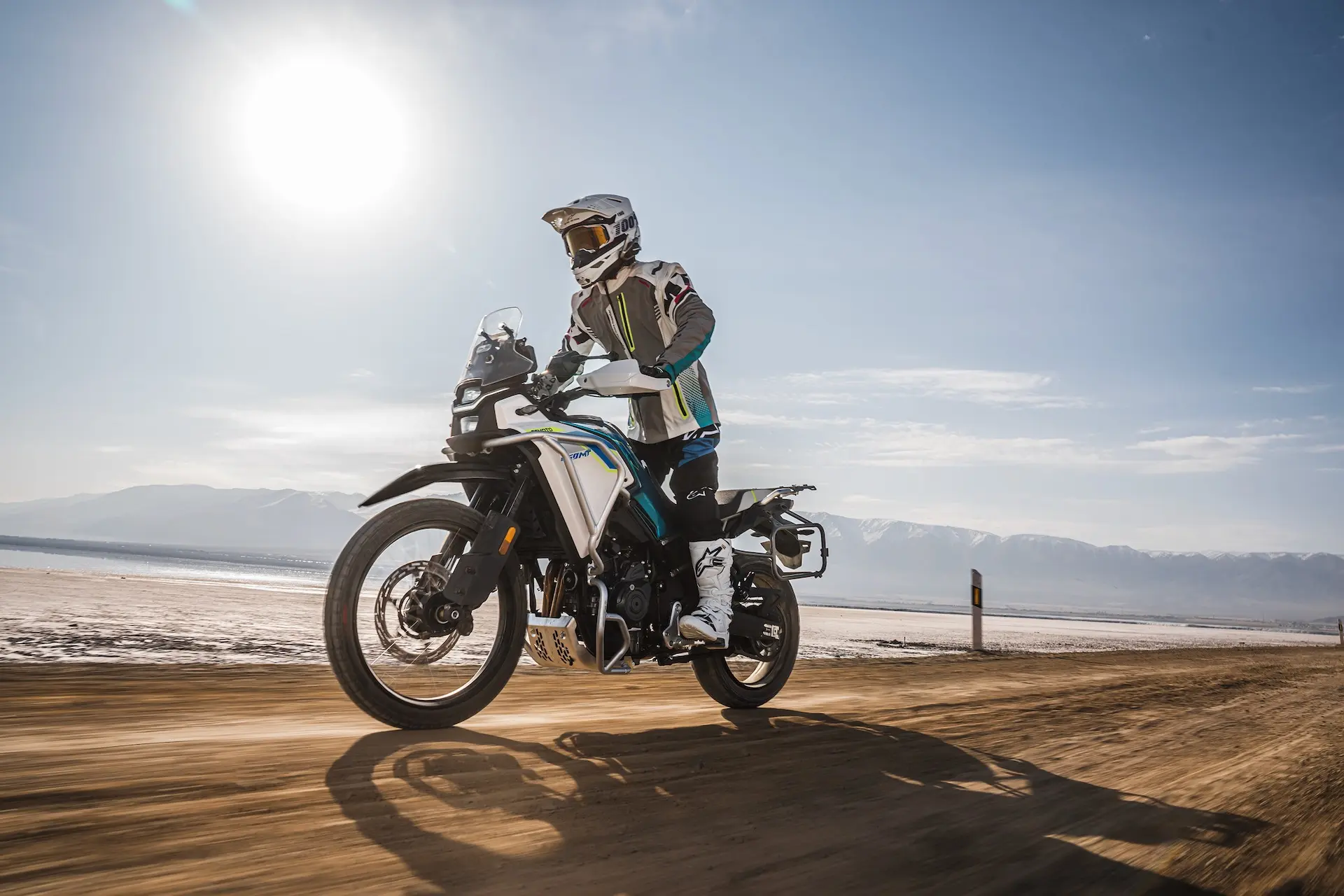 A person wearing protective gear rides an A2 Motorbikes UK adventure motorbike on a dirt road, with mountains and the sun in the background.